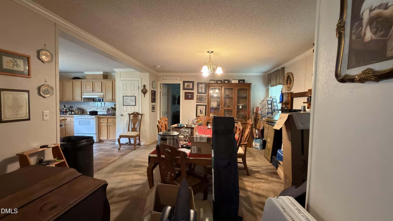 A cluttered dining and living area with wooden furniture, boxes, framed pictures on the walls, and a kitchen in the background. Light fixtures hang from the ceiling and various items are scattered around the room.