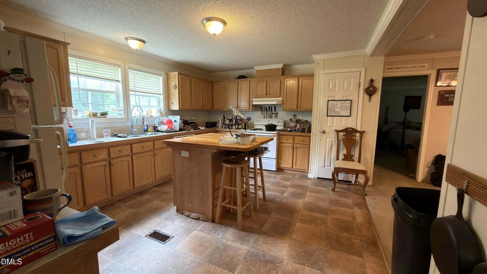 A spacious kitchen with wooden cabinets, an island in the center, various kitchen items on counters, a double sink under windows, a stove, and a chair by a doorway leading to another room.