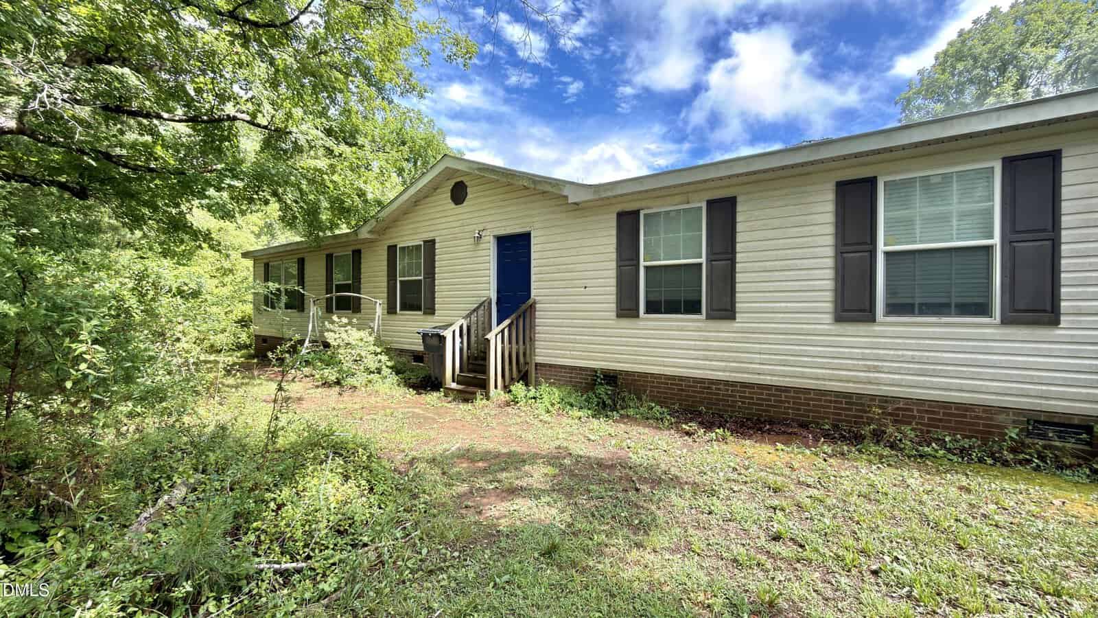Single-story manufactured home at 4552 Gray Rock Road in Kittrell with beige siding, black shutters, and a blue front door. The house is surrounded by overgrown grass and trees, with a small porch and steps leading to the entrance under a partly cloudy sky.