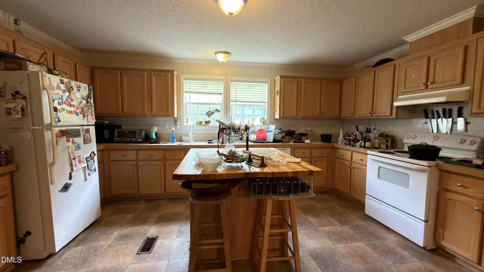 Spacious kitchen with wooden cabinets, white appliances, a center island with stools, and various items on counters. Natural light comes through double windows above the sink.