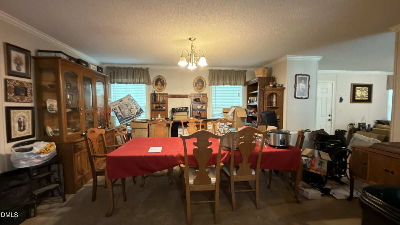 Dining room with a red tablecloth, wooden chairs, and various household items cluttering the space. Cabinets, wall art, and windows with blinds in the background add to the cozy, lived-in atmosphere.