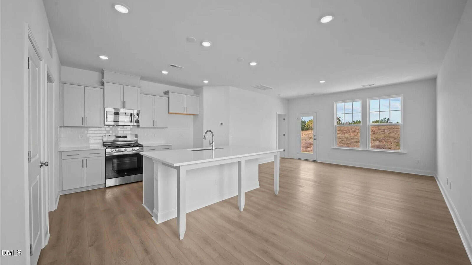 A modern, bright kitchen with white cabinets, stainless steel appliances, and an island overlooks an open living area with large windows and light wood floors.