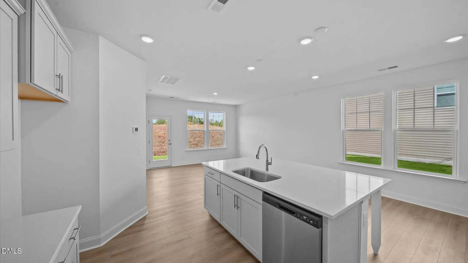 Modern, bright kitchen with white cabinets, island with sink, stainless steel dishwasher, light wood floors, and large windows letting in natural light.