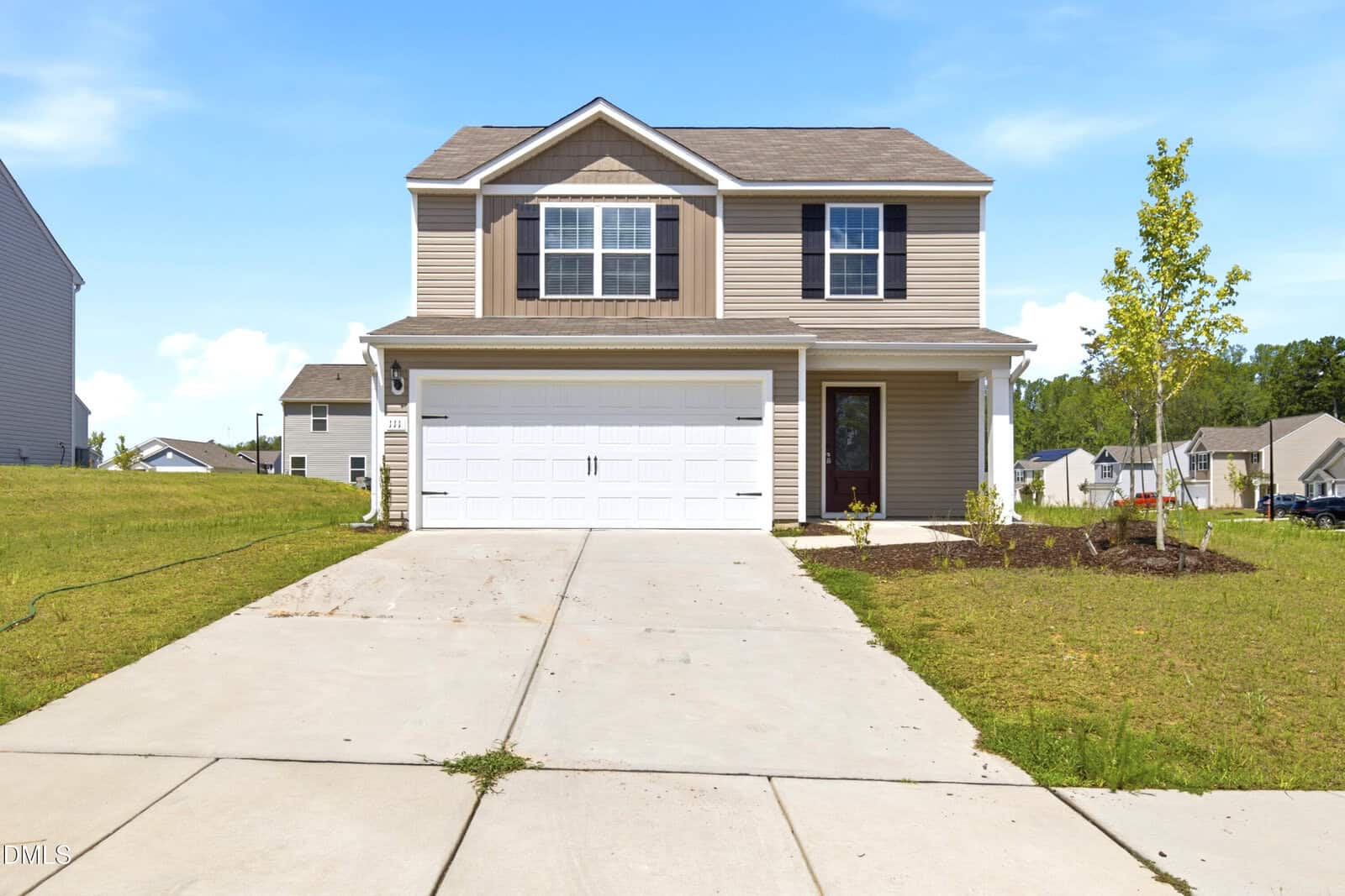 A two-story beige house with white trim and a double garage sits on a concrete driveway. The lawn is partially landscaped with a small tree and neighborhood houses are visible in the background under a blue sky.