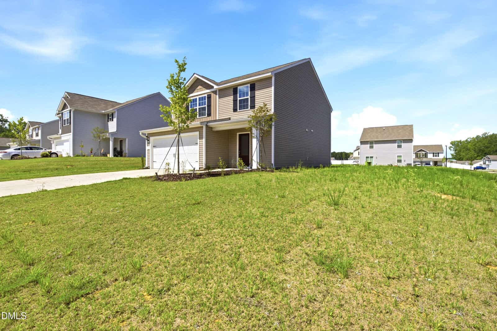 Two-story suburban house with two-car garage, beige siding, and young tree in the front yard. The house sits on a grassy, slightly sloped lawn in a residential neighborhood under a blue sky.