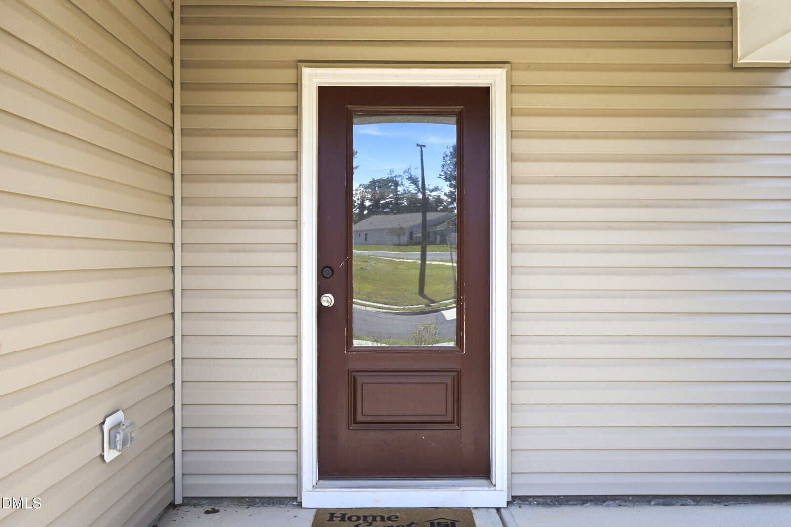 A brown door with a glass window is set in tan siding. A doormat with faint text sits at the threshold, and a neighborhood street is reflected in the glass.