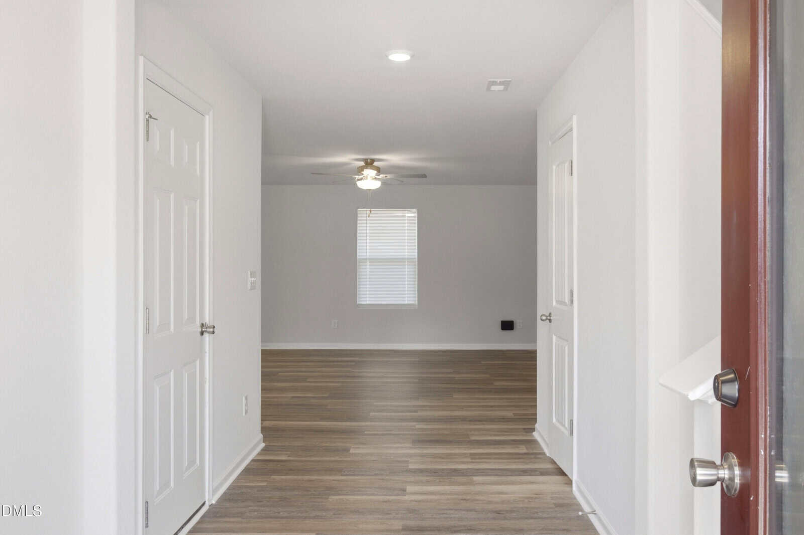 View from an open door into a hallway with light wood floors, white walls, and two closed doors, leading to a bright room with a window and a ceiling fan.