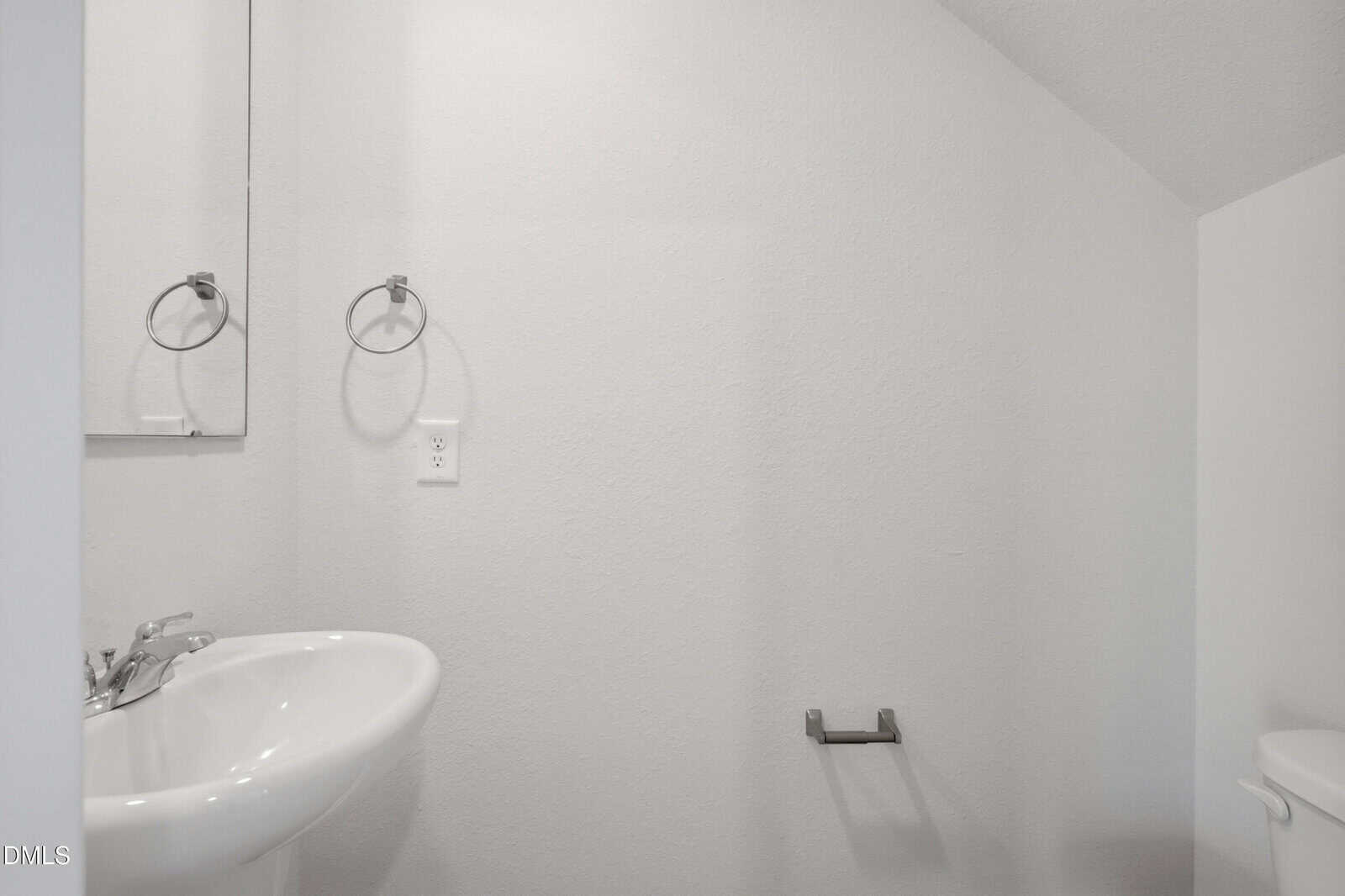 Minimalist bathroom with white walls, a small sink, faucet, mirror, towel ring, empty toilet paper holder, and a partial view of a toilet. The lighting is bright and the decor is simple.