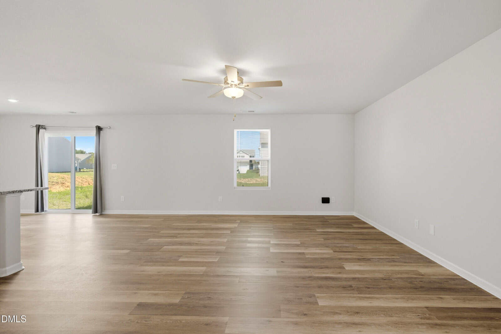 A spacious, empty room with light wood flooring, white walls, a ceiling fan with lights, a window, sliding glass doors, and ample natural light.