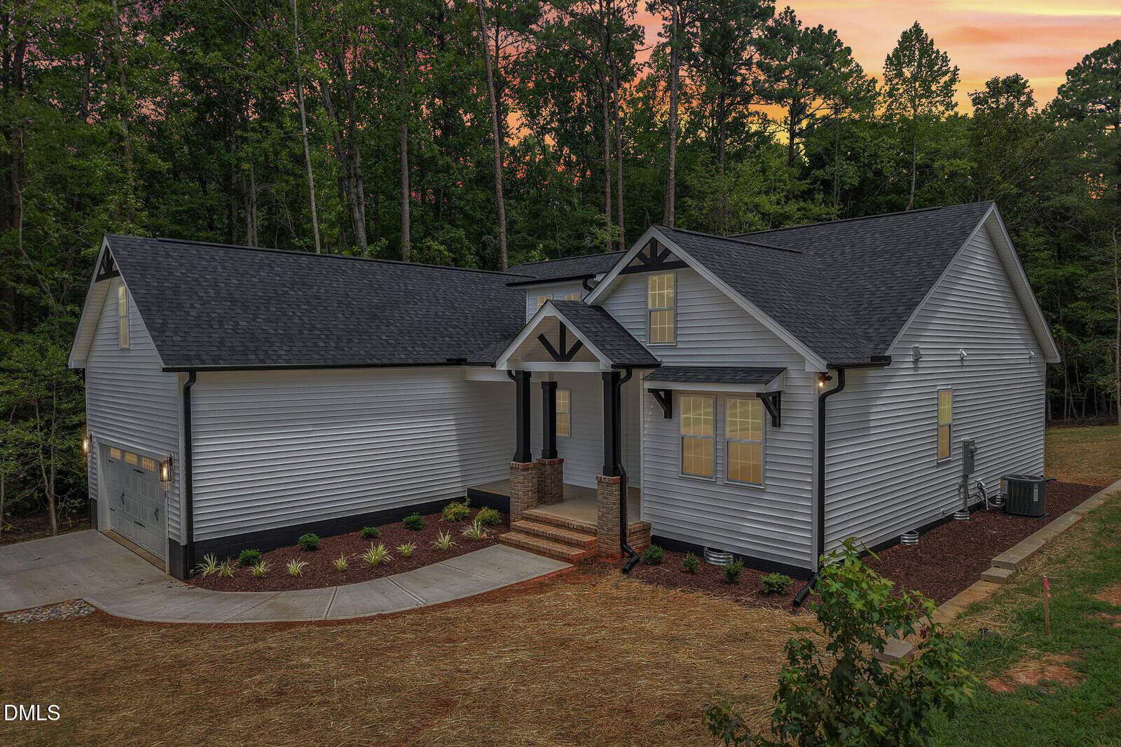A modern single-story gray house with white trim, black roof, covered front porch, and attached garage sits at 1316 Sagamore Drive in Louisburg, surrounded by trees and a landscaped yard at sunset.