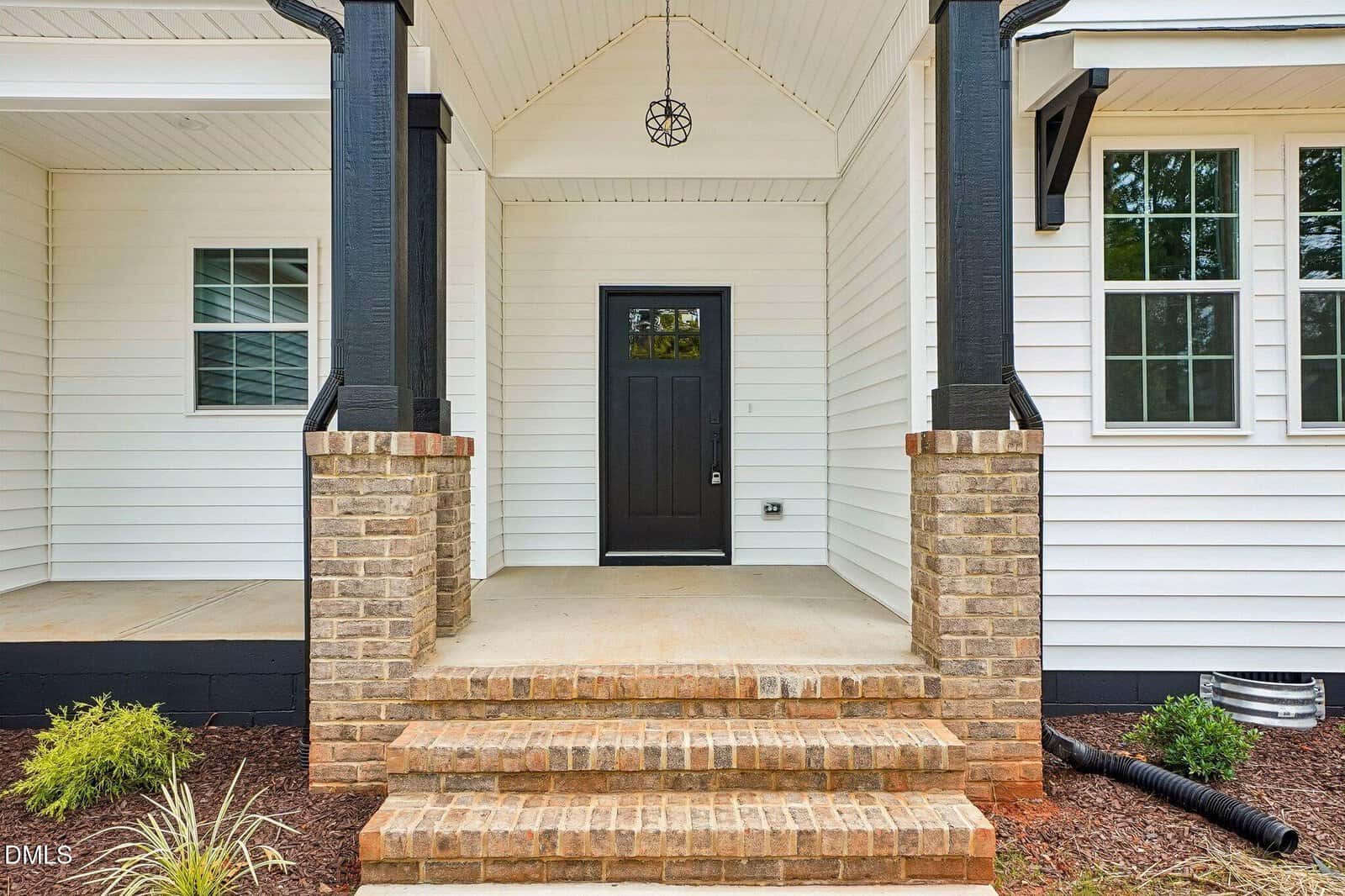 A front porch with brick steps, black columns, and a black front door on a white house. There are windows on both sides and landscaping with small plants.