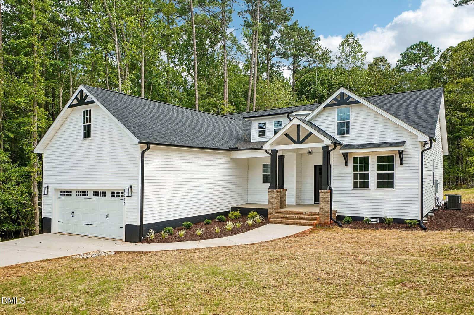 A modern white house with black trim, double garage, gabled roof, covered porch with columns, and a landscaped front yard, set against a backdrop of tall trees and blue sky.