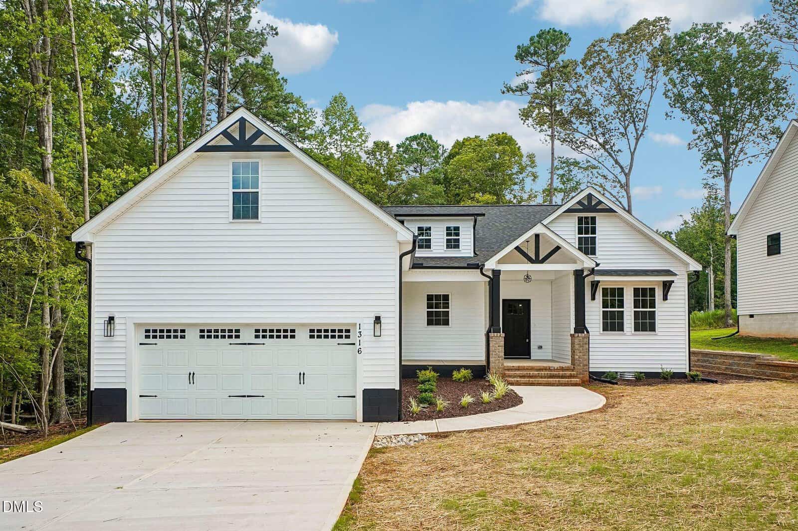 A white two-story house with black trim, a double garage, front porch, and gabled roof sits on a spacious lot surrounded by trees under a partly cloudy sky.