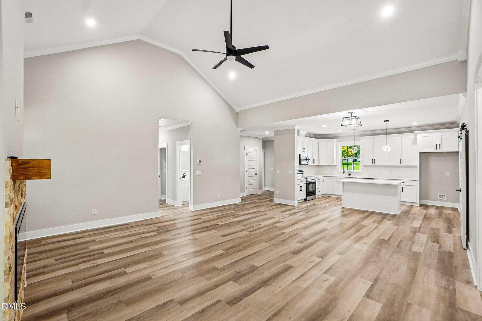 Open-concept living area with light wood floors, white walls, vaulted ceiling, ceiling fan, modern kitchen with island, white cabinets, stainless steel appliances, and large window with green view.