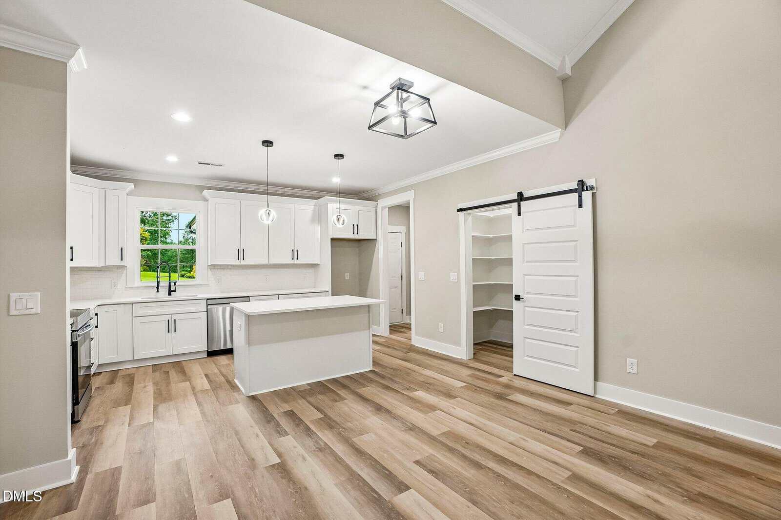 Modern kitchen with white cabinets, an island, pendant lights, and light wood floors. A sliding barn door opens to a pantry. A window over the sink lets in natural light. Walls are light gray, and the ceiling has crown molding.