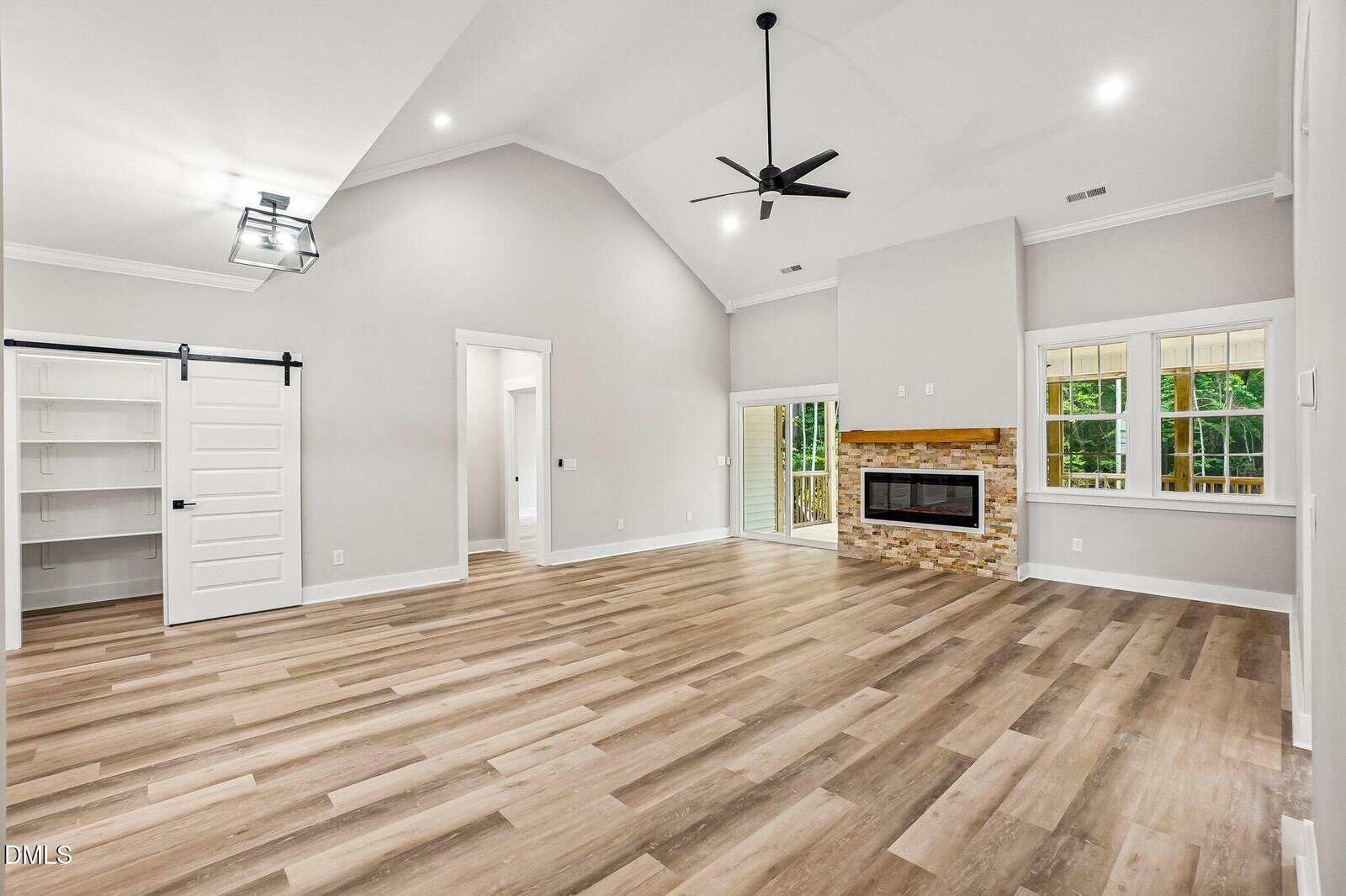 Spacious living room with vaulted ceiling, light wood flooring, stone fireplace, ceiling fan, sliding barn door to closet, and large windows overlooking greenery.