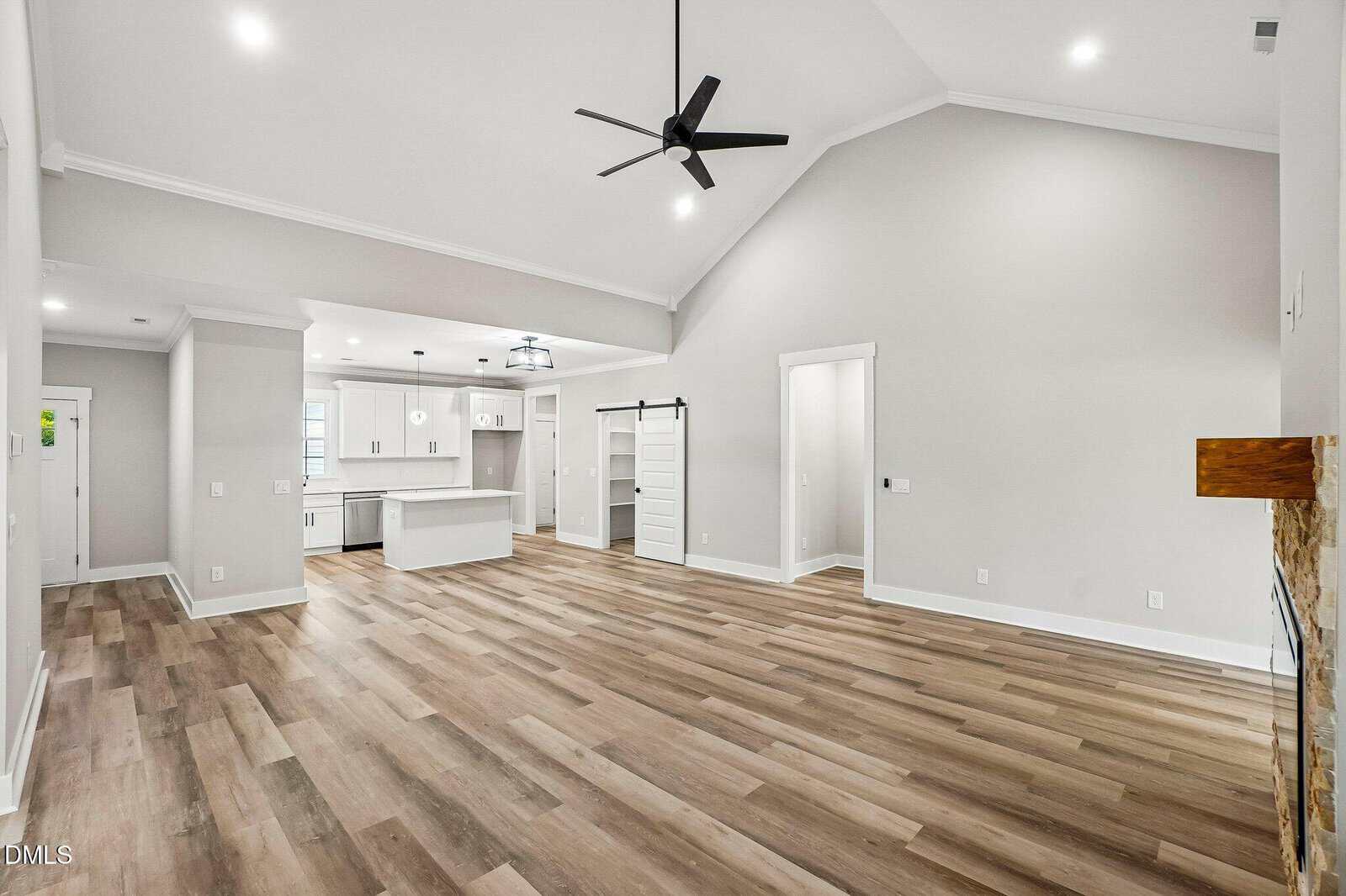 Spacious modern living area with light wood flooring, vaulted ceiling, black ceiling fan, white walls, and open kitchen featuring white cabinets and an island. A sliding barn door and a fireplace are also visible.