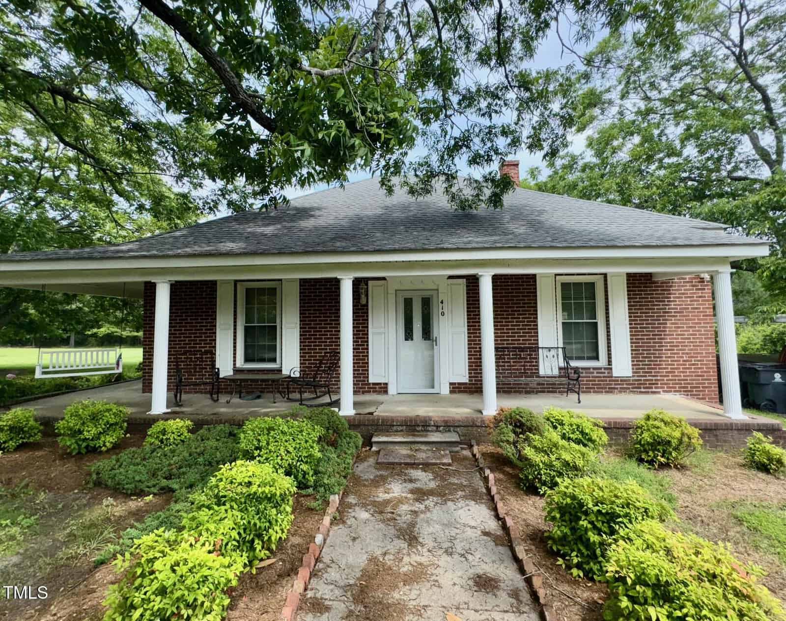 A brick house on 410 High Street in Norlina, featuring a large covered porch with white columns and two benches. Green bushes line the front walkway, while leafy trees provide a picturesque backdrop.