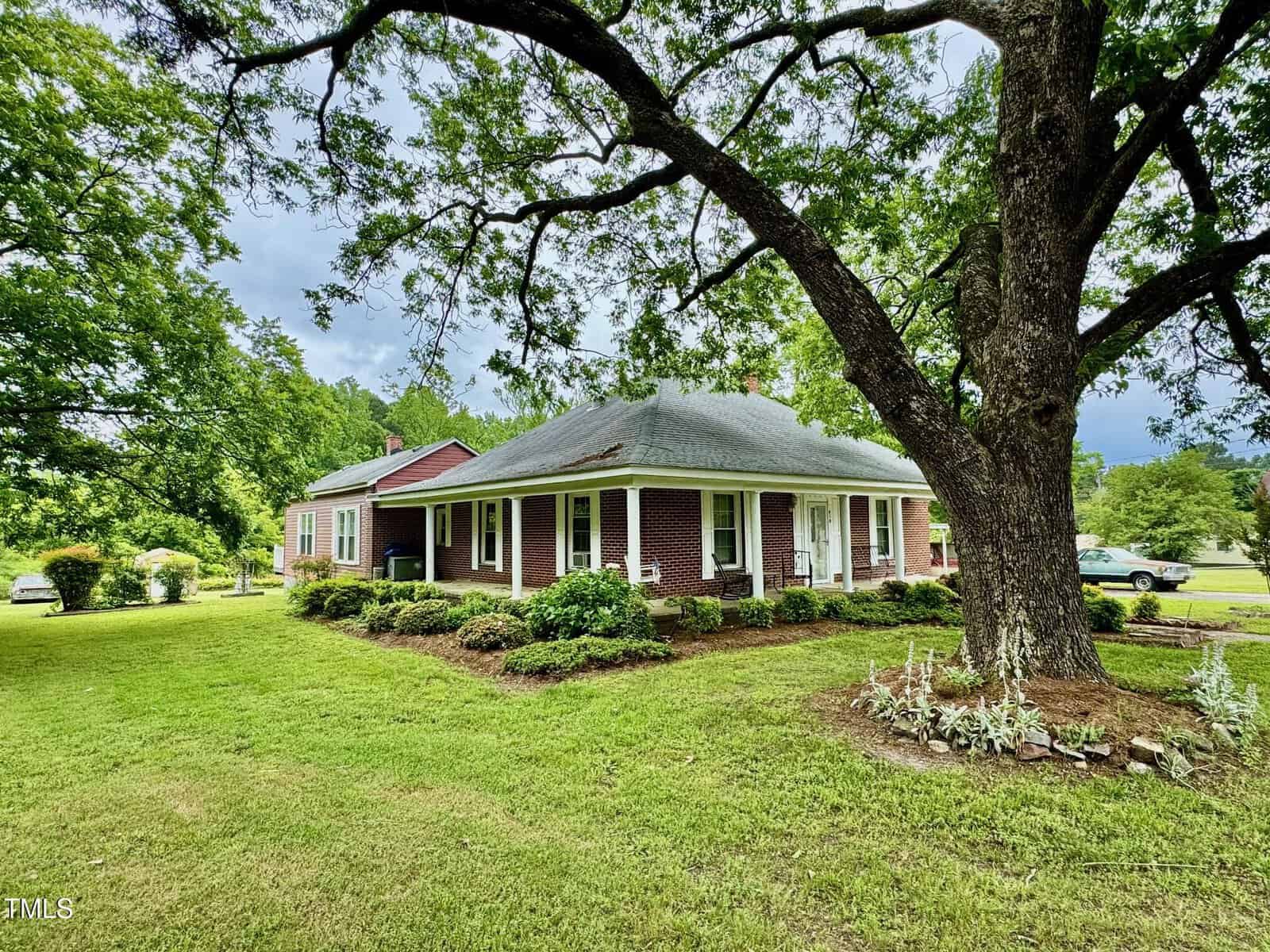 A red-brick house with white trim sits on a grassy lawn, shaded by a large tree. Bushes and plants line the house, and a car is visible in the background. The sky is partly cloudy.