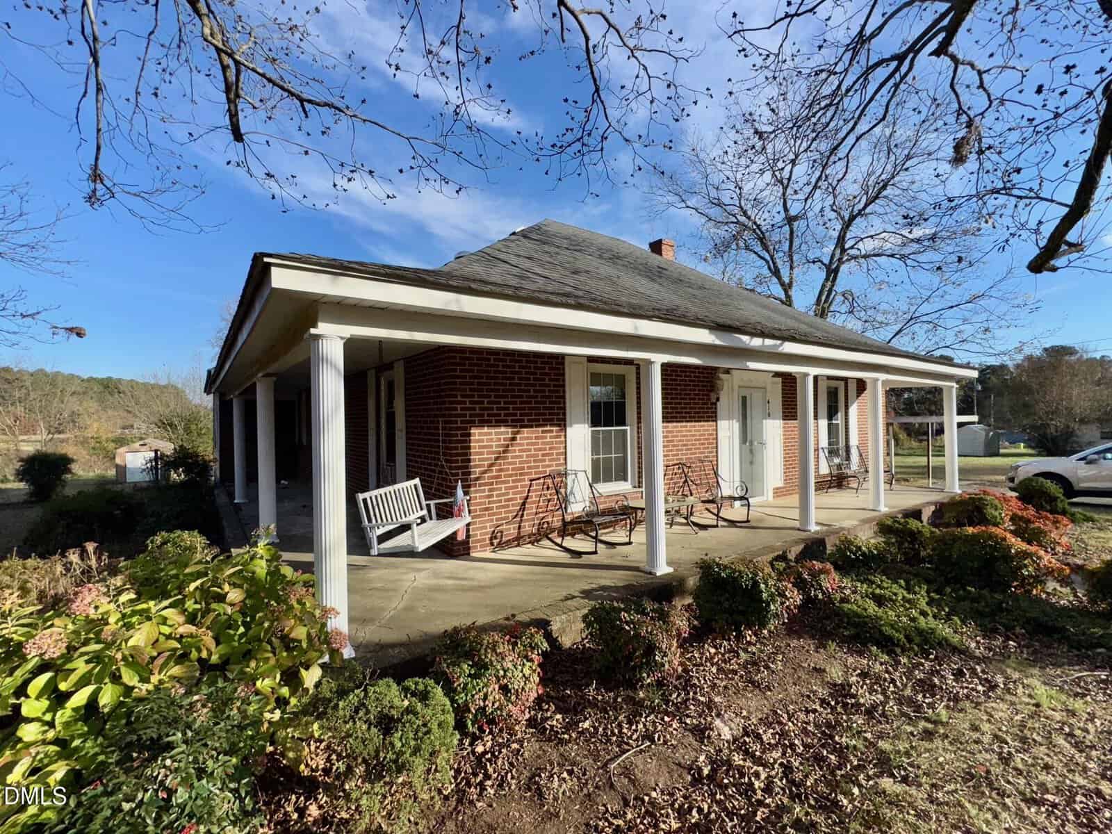 A red brick house with a large covered porch, white columns, porch swings, and rocking chairs, surrounded by shrubs and trees under a blue sky.