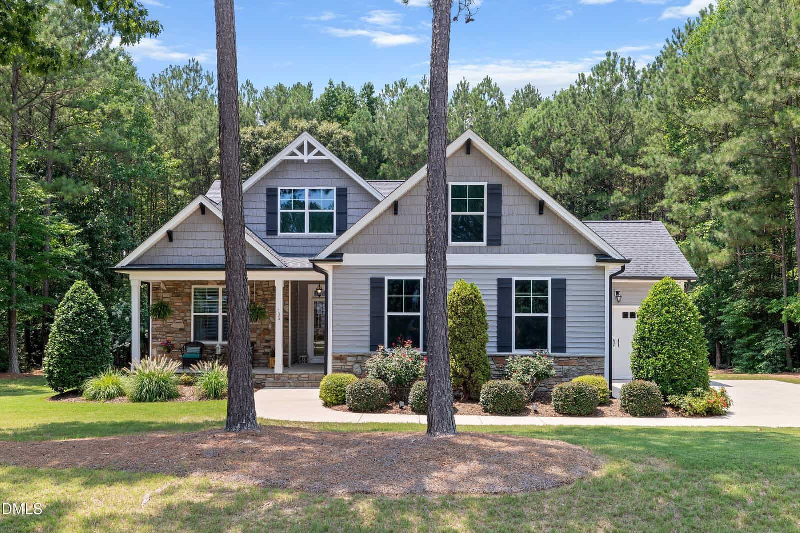 A modern two-story house with gray siding, stone accents, and a front porch, 105 Carriden Drive in Youngsville is surrounded by neatly trimmed bushes, trees, and a wooded background. A paved driveway leads to a side garage.
