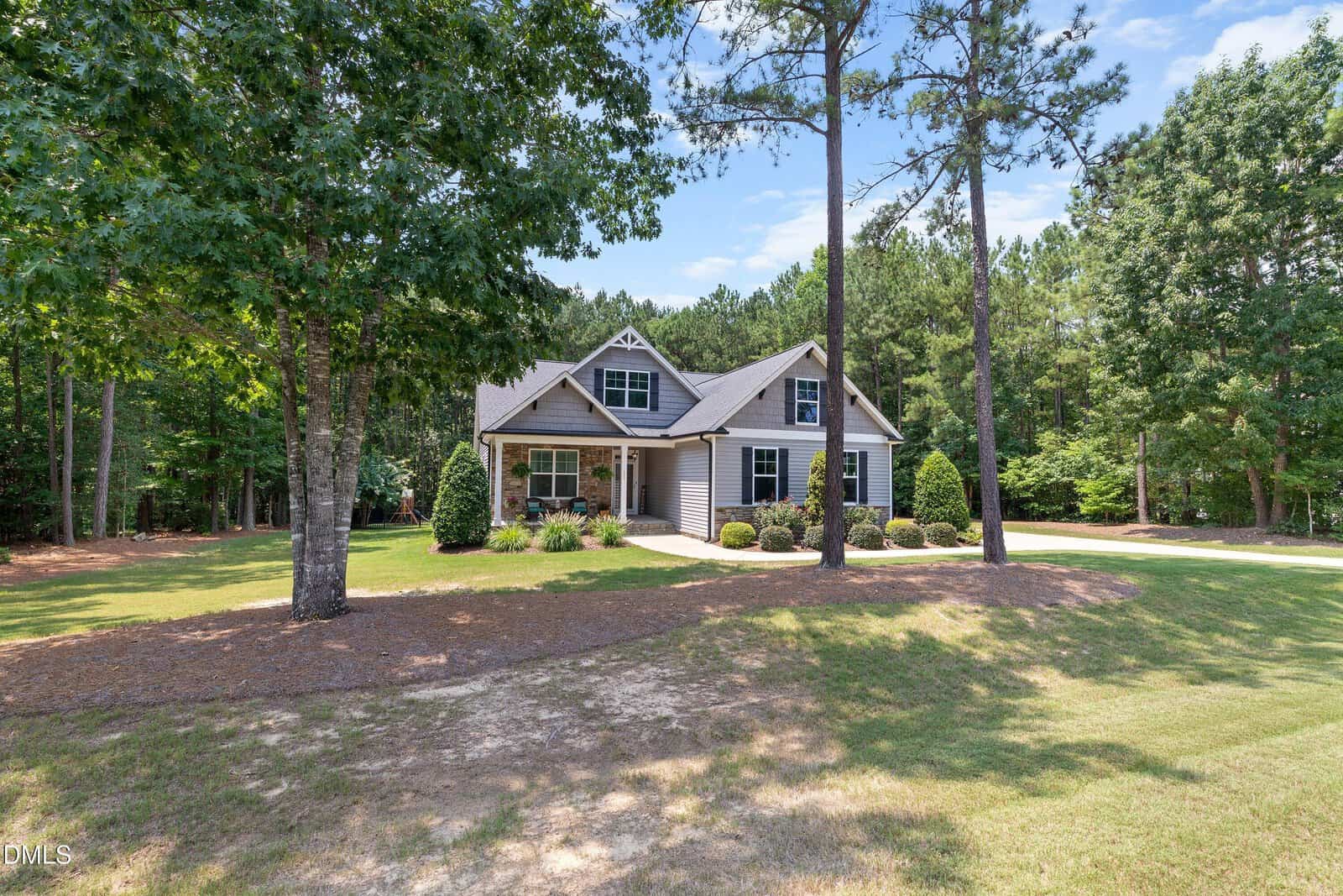 A two-story house with gray siding and a covered porch sits on a large, grassy lot surrounded by tall trees under a blue sky. A curved driveway leads to the home, and landscaped bushes line the front.