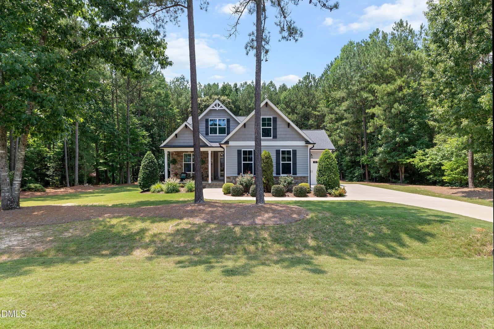 A single-story house with a gabled roof and front porch sits among tall trees, with a curved driveway and well-kept lawn in a wooded setting.