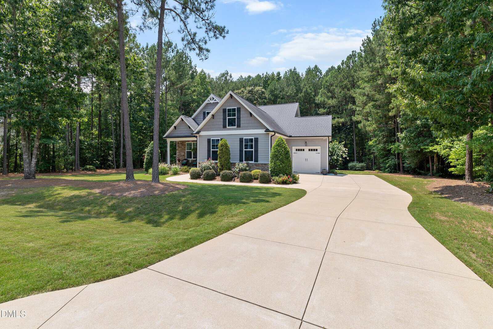 A gray house with a gabled roof, front porch, and single-car garage sits surrounded by trees. A long, curved driveway leads from the road to the garage. The lawn is neatly maintained.