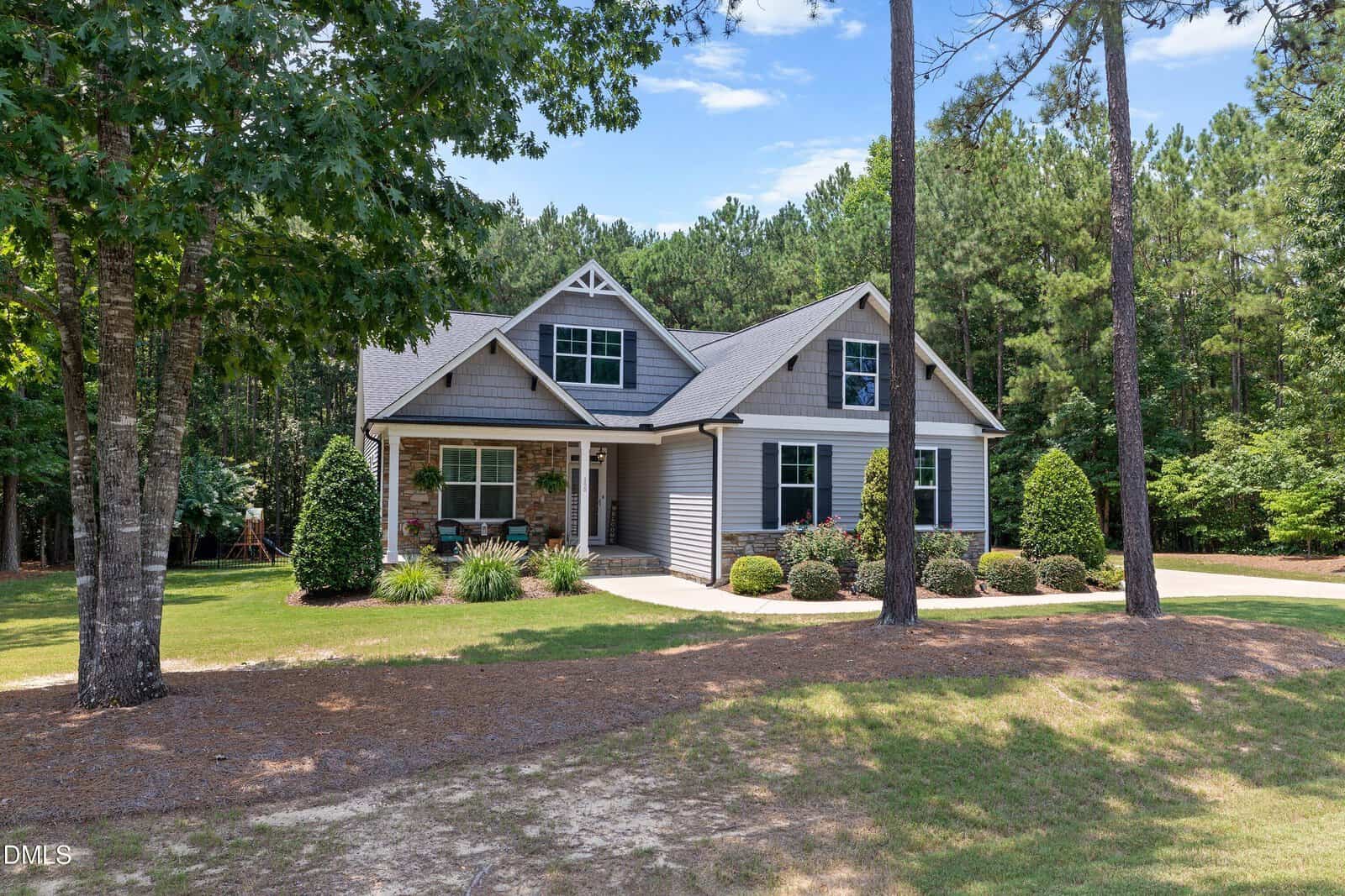 A gray two-story house with white trim sits among tall trees, featuring a covered front porch, manicured shrubs, and a spacious, grassy yard under a clear blue sky.