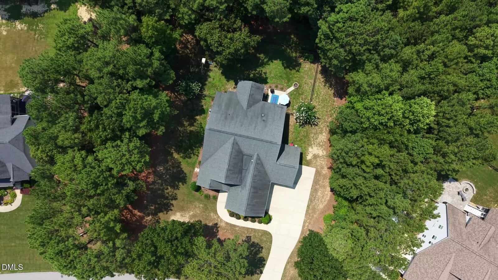 Aerial view of a house with a dark roof, a driveway, a small backyard pool, and surrounding trees providing dense green coverage.