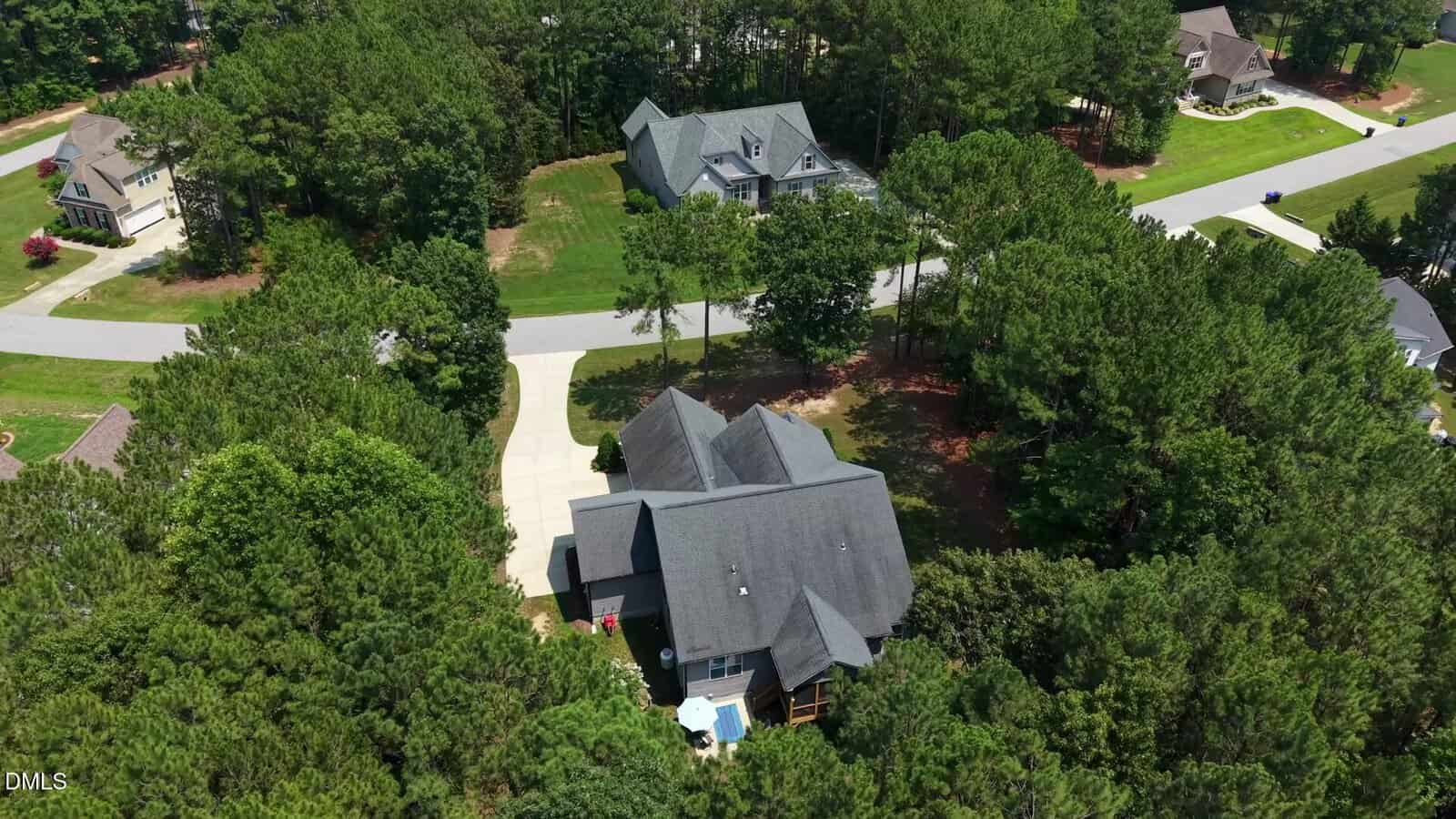 Aerial view of a house surrounded by trees, with a long driveway leading to a street. Neighboring houses and more trees are visible in the background.