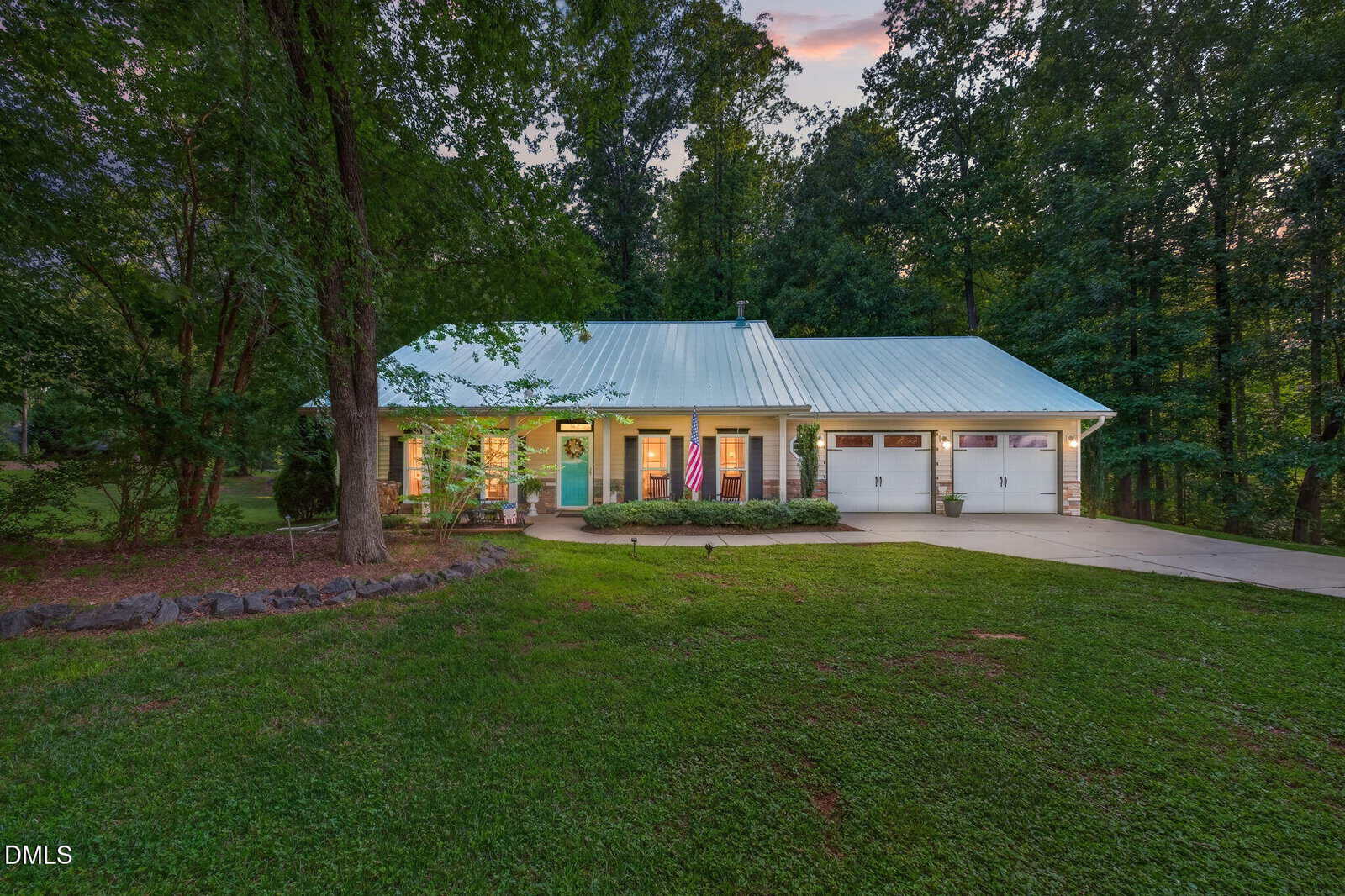 A single-story house with a metal roof, front porch, and attached garage sits surrounded by trees and grass at dusk on 137 Sacred Fire Road in Louisburg. An American flag hangs by the front door.