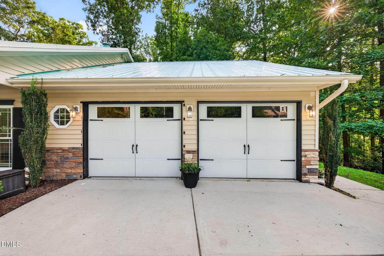 Two white garage doors with windows, black hardware, and a central potted plant. Beige house with stone accents, surrounded by green trees and sunlight filtering through foliage.