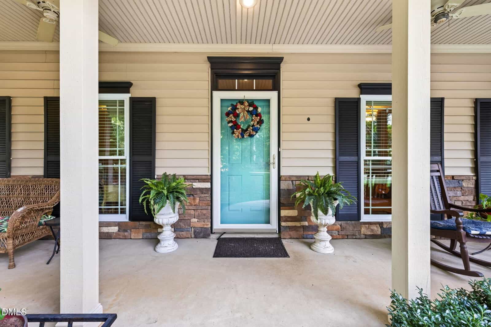 A front porch with a light blue door decorated with a wreath, flanked by two potted plants, white columns, rocking chairs, and windows with black shutters on either side.
