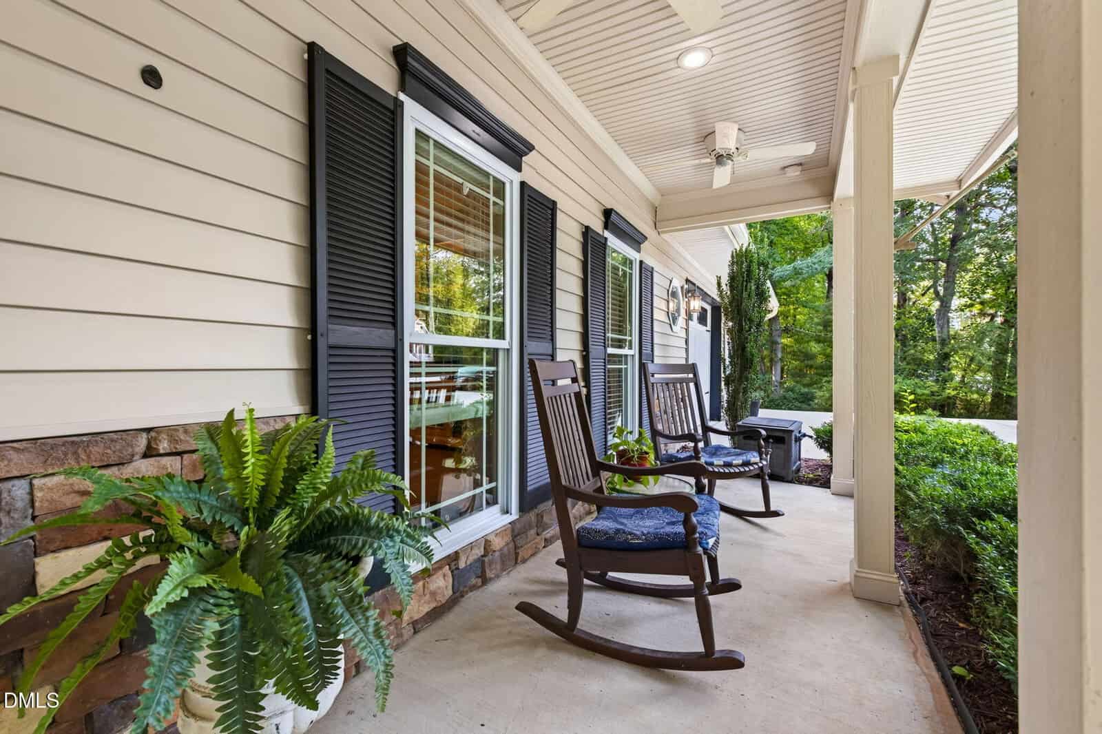 Front porch with two wooden rocking chairs with blue cushions, a small table, potted fern, and black shutters on windows. The porch overlooks a green, leafy yard.