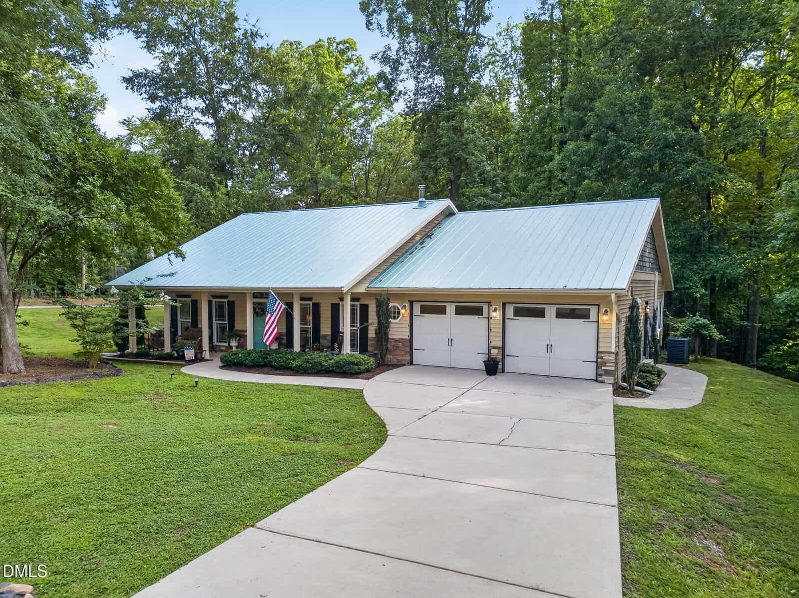 A single-story house with a metal roof, covered porch, two-car garage, an American flag hanging by the entrance, surrounded by trees and a neatly manicured lawn with a curved driveway.