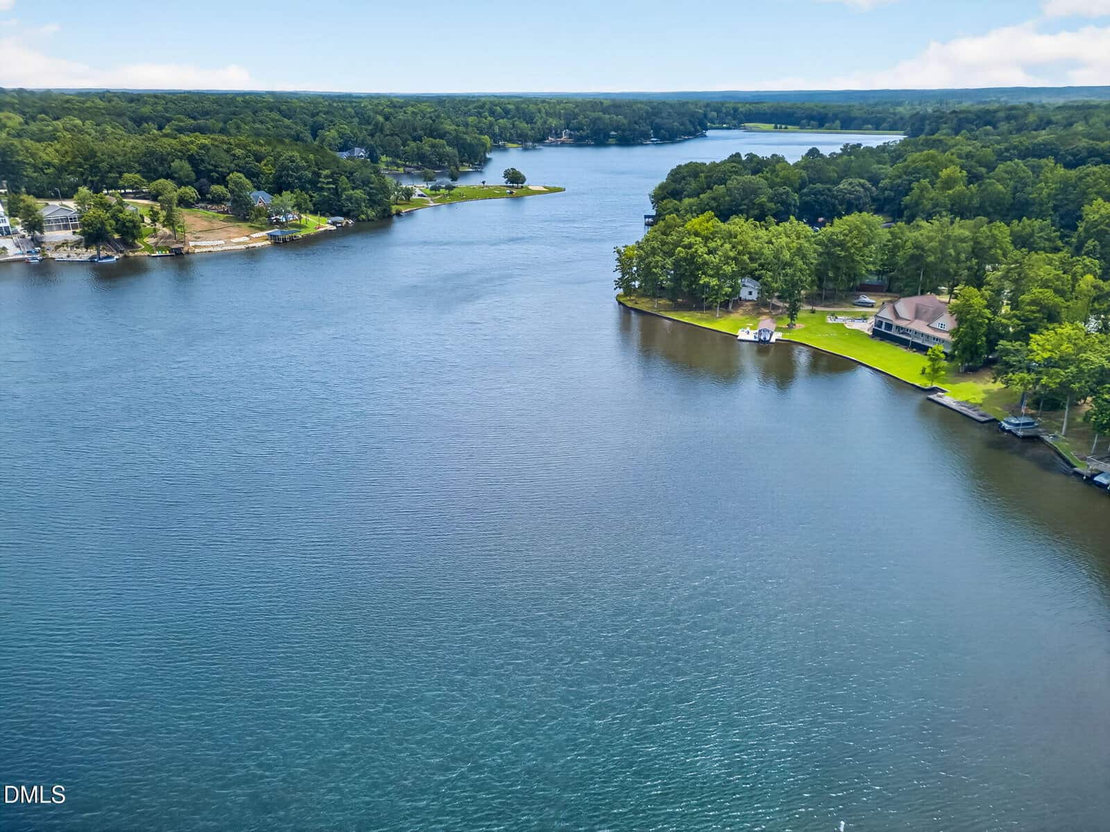 Aerial view of a peaceful lake surrounded by trees and houses, with calm blue water and partly cloudy skies in the background.