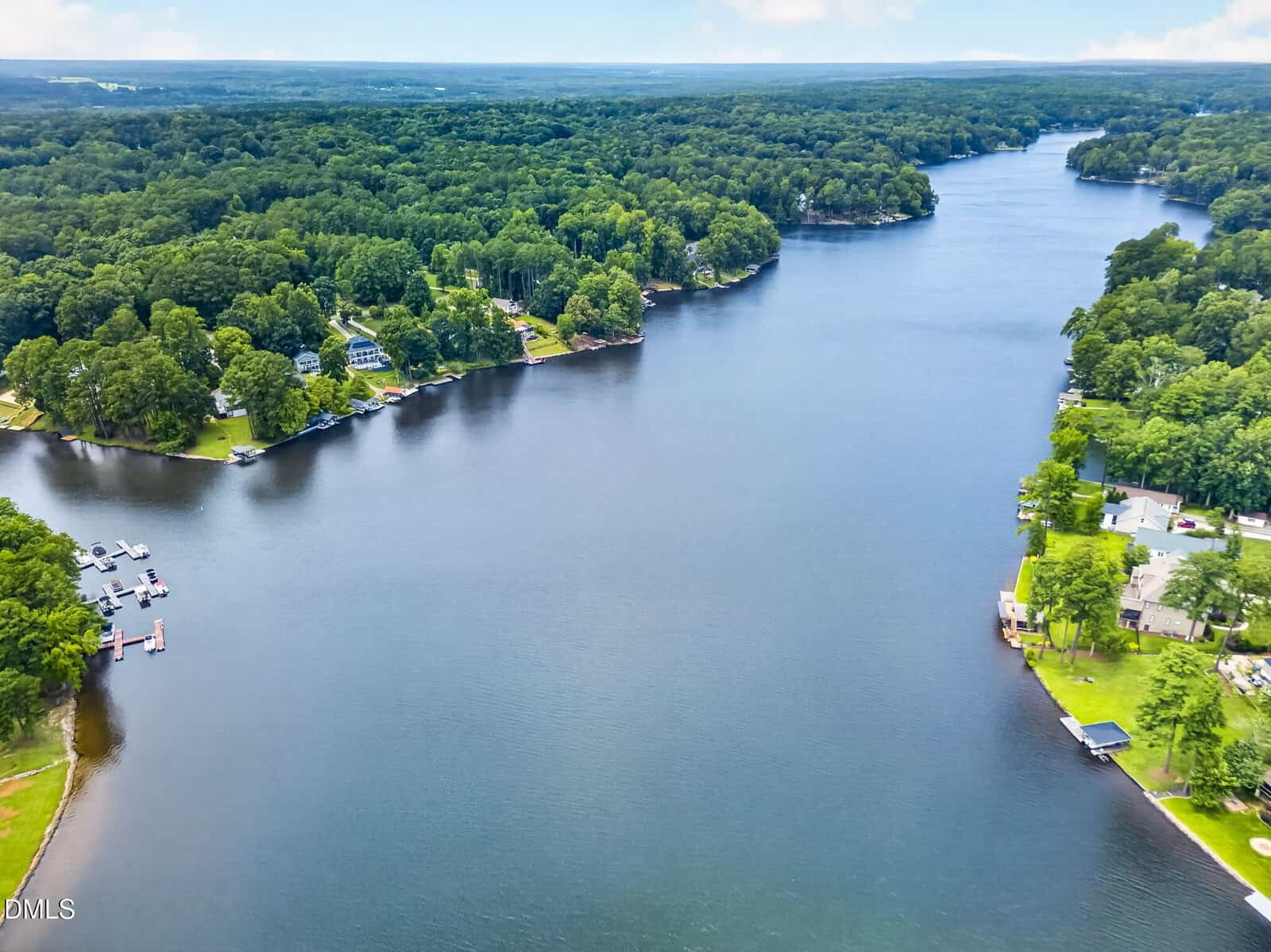 Aerial view of a wide river or lake with tree-lined shores and scattered houses, docks, and boats along the water’s edge, surrounded by dense green forest under a partly cloudy sky.