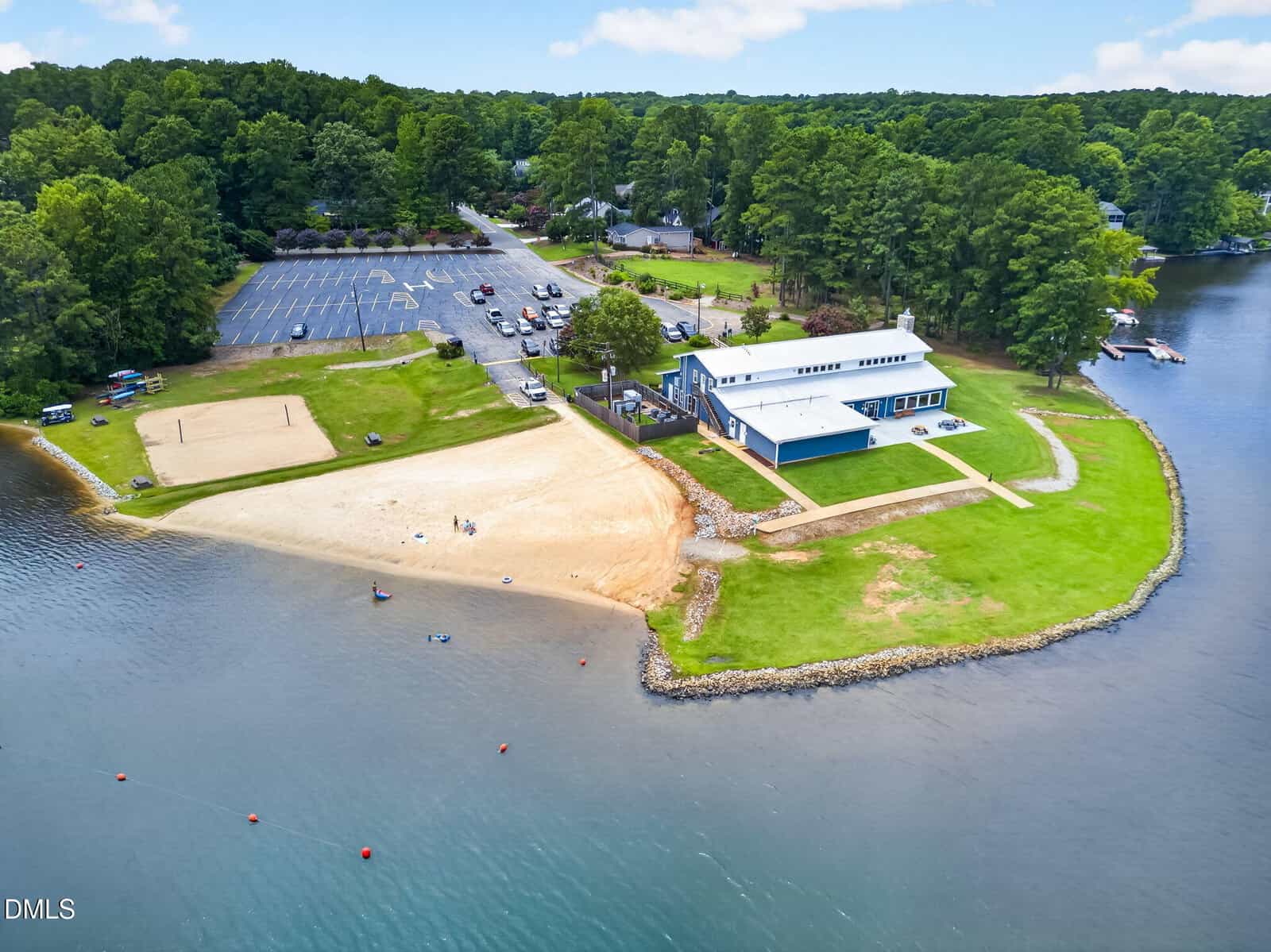 Aerial view of a lakeside recreation area with a sandy beach, large blue building, grassy lawn, parking lot, volleyball court, and forested area in the background.