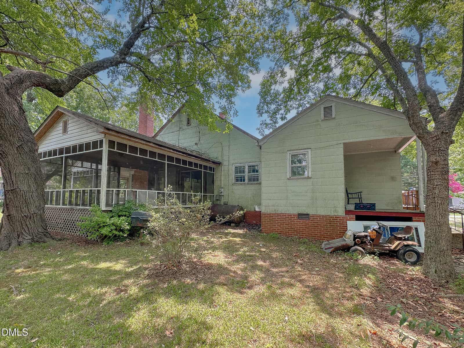 A light green house with a screened porch, carport, and large trees providing shade. Old toys and a small vehicle are in the yard, and sunlight filters through the leaves onto the grass.