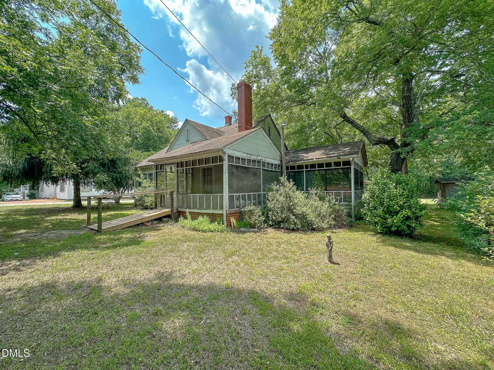 A quaint house with a screened porch, green siding, and a brick chimney sits among tall trees and lush greenery under a partly cloudy sky.