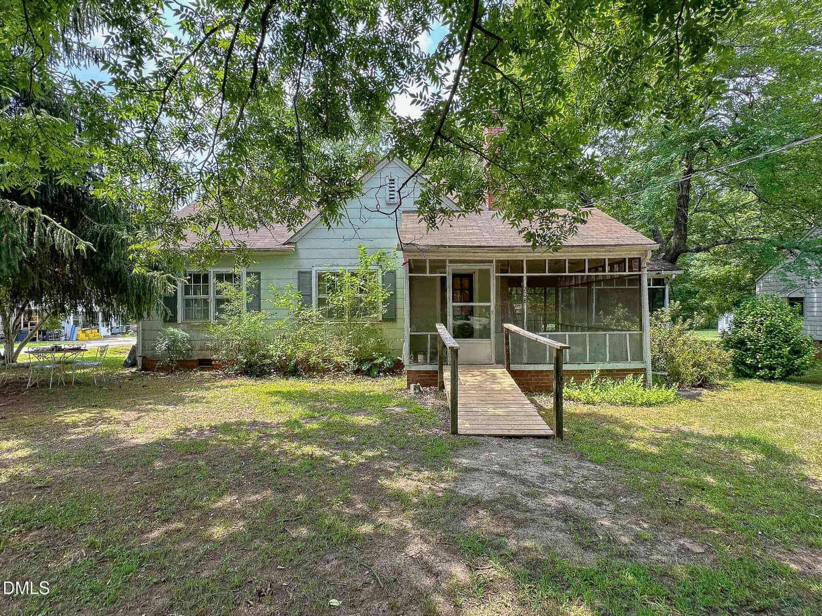 A light green house with a brown roof, screened front porch, and wooden ramp sits among trees and grass on a sunny day in Norlina at 208 Elm Street.