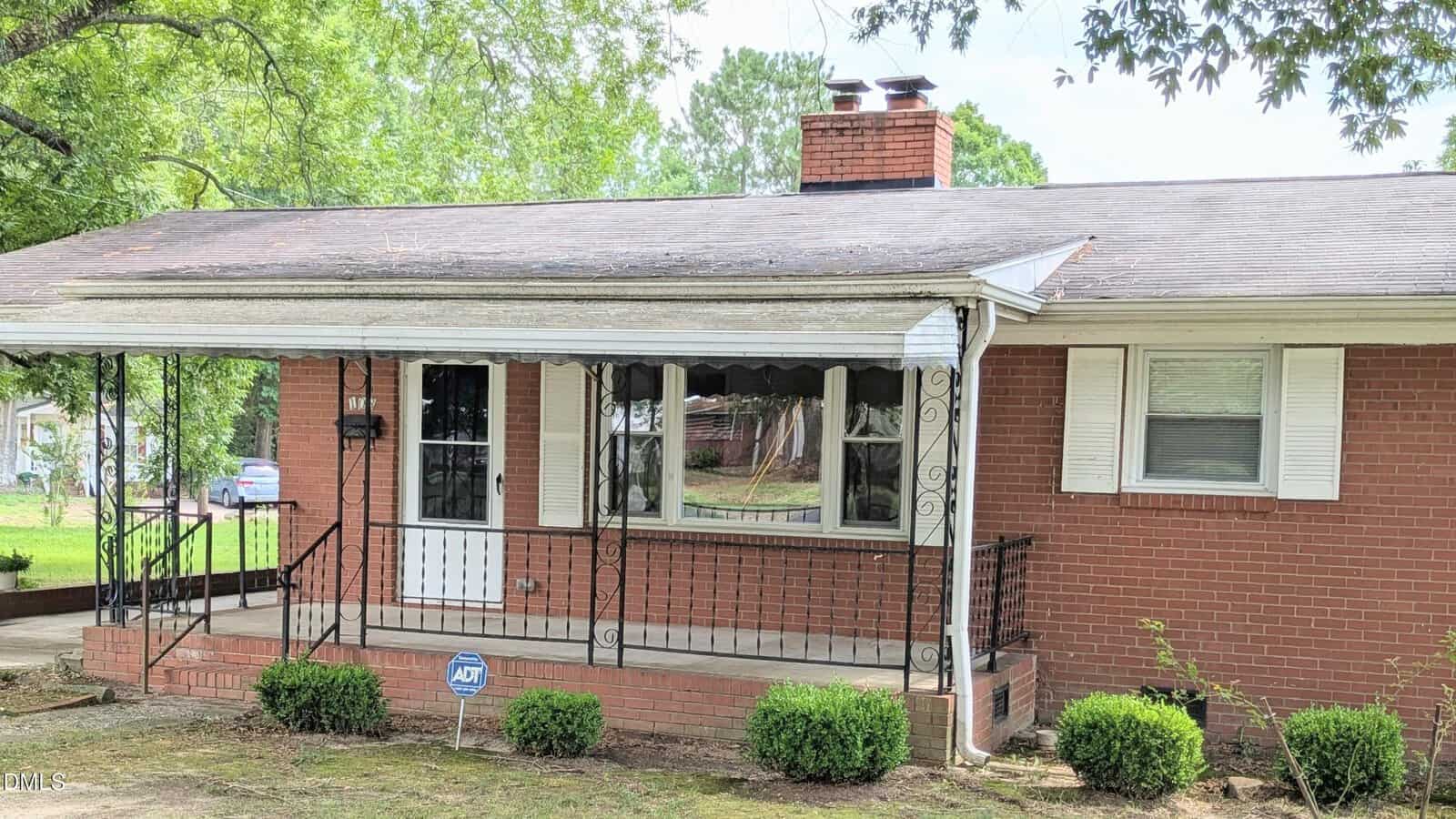 Single-story red brick house with a small covered front porch, white trim, black metal railing, a chimney, and an ADT security sign in front. Bushes line the edge of the porch, and trees surround the yard.