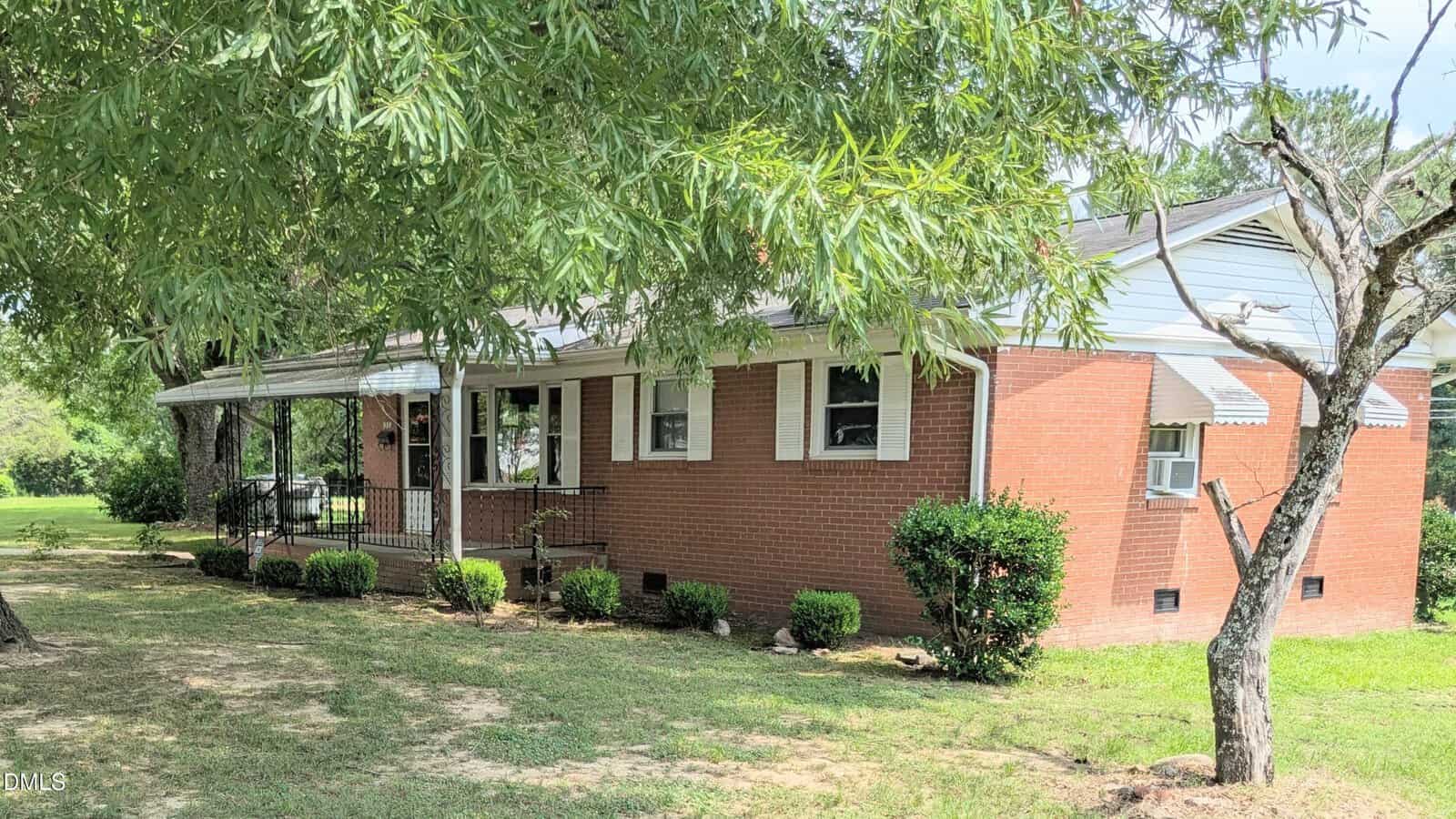 A single-story red brick house with white trim, a covered front porch, and green shrubs. The yard is grassy with some trees providing shade in the foreground.