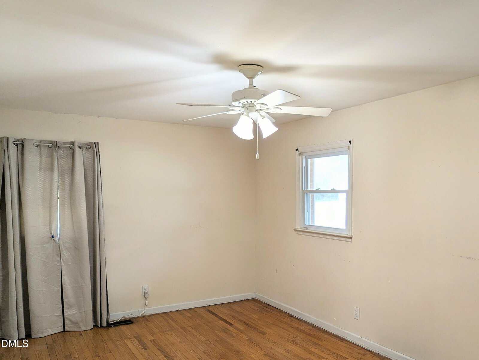 A simple, empty room with light-colored walls, a hardwood floor, a ceiling fan with lights, a window with blinds, and a curtain covering another window.
