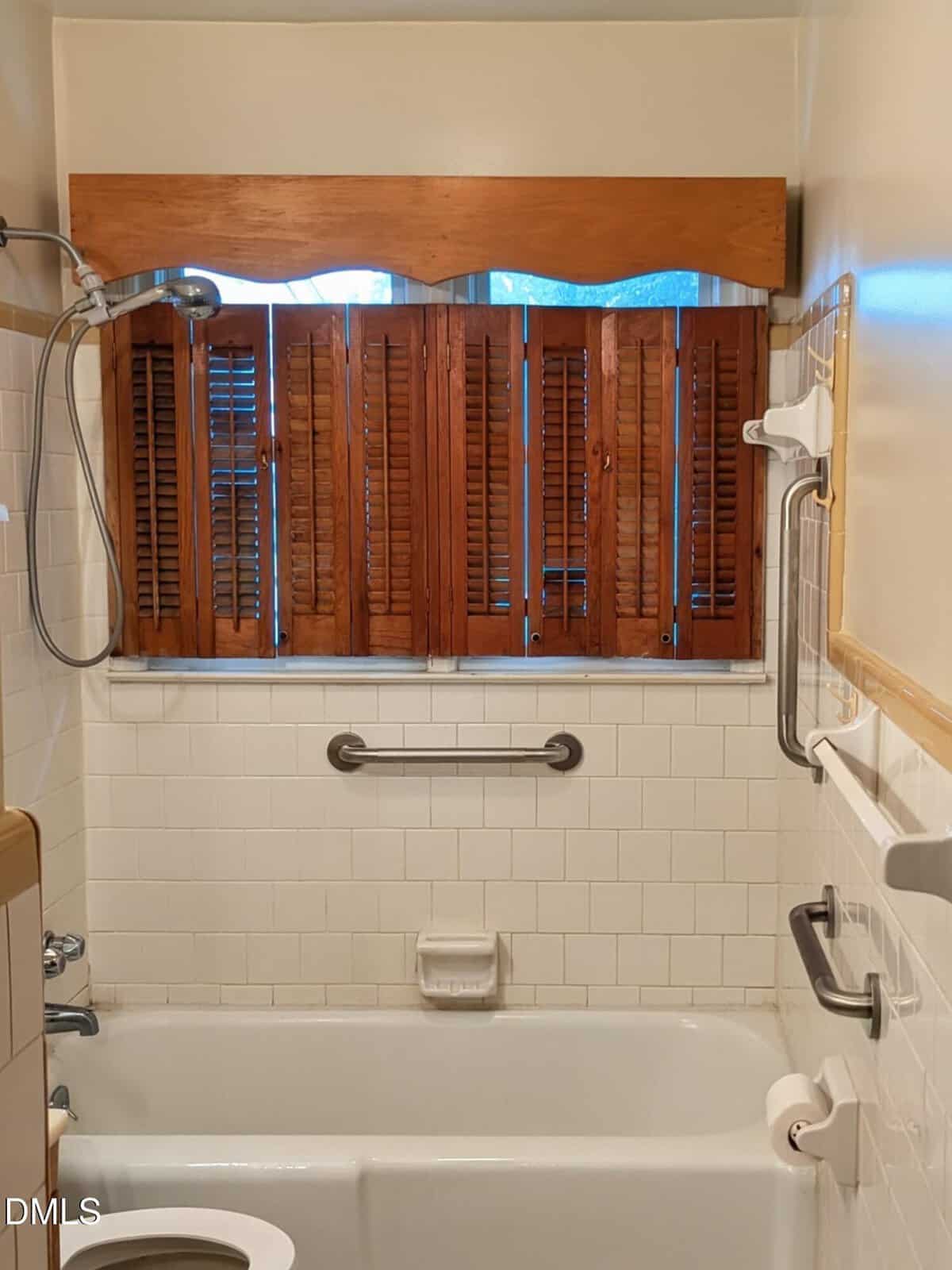 Bathroom with a white tiled shower and bathtub, brown wooden shutters on the window, grab bars on the walls, a soap holder, and a toilet paper holder visible on the right.