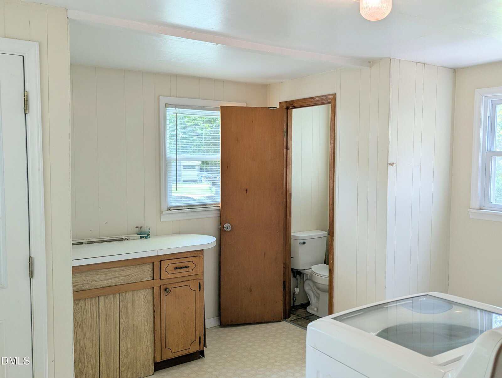 A bright laundry room with a washer, a wooden cabinet with a sink, and a door leading to a small bathroom with a toilet. White walls and windows let in natural light.