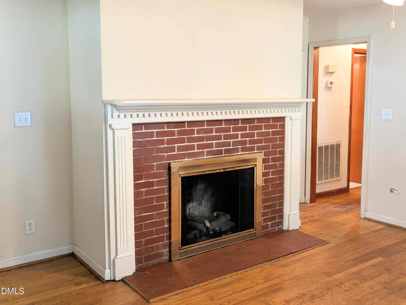 A red brick fireplace with a white wood mantel sits against a beige wall on a hardwood floor. The fireplace has a black firebox and a gold metal trim around the opening.