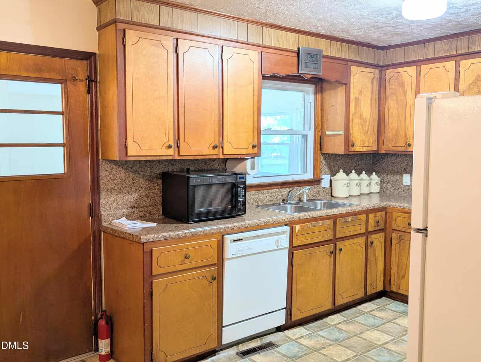 Wood-paneled kitchen with brown cabinets, a white dishwasher, sink, countertop microwave, white refrigerator, and a window above the sink. Four canisters and a decorative sign sit on the counter.