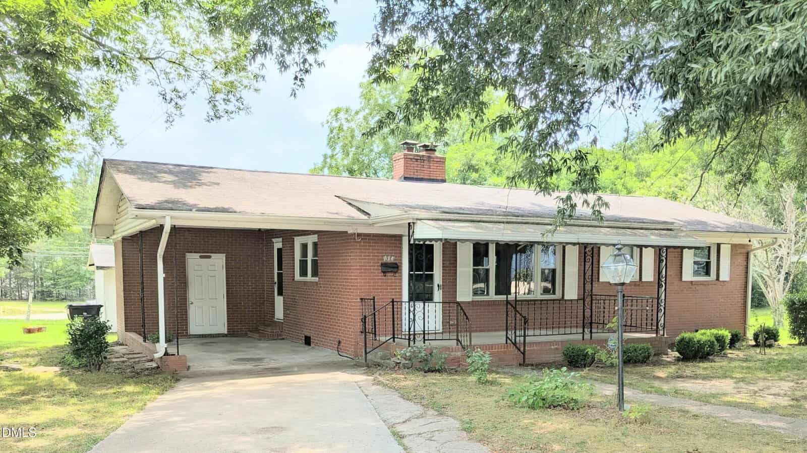 Single-story brick house at 103 Hayes Street, Louisburg, featuring a covered front porch with black railing, chimney, driveway, and a white side door. The yard has some bushes and is shaded by trees, with sunlight filtering through the leaves.
